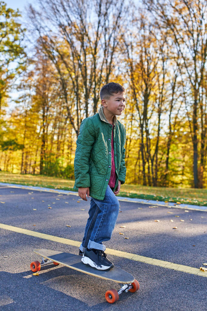 preteen african american boy in outerwear and jeans riding penny board, autumn park, fall season - Фото, изображение