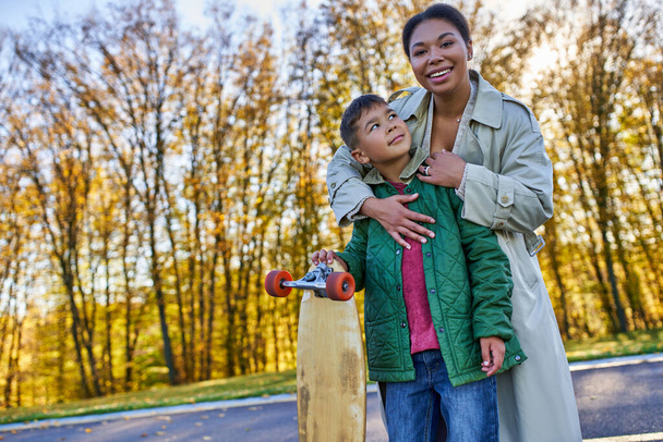 happy african american woman hugging son with penny board, autumn, fall season, motherly love, park - Фото, изображение