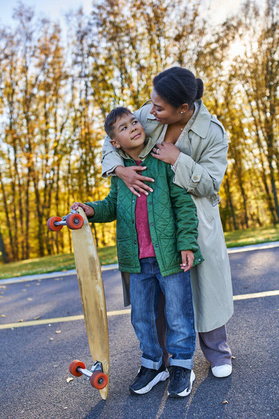 african american mother hugging son with penny board, autumn, fall season, motherly love, smile - Foto, Imagen