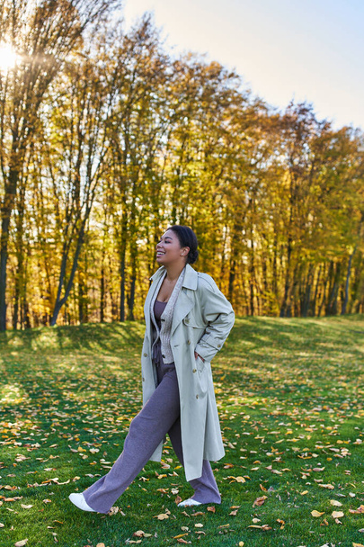 mujer afroamericana feliz en gabardina caminando sobre hierba con hojas caídas, otoño, moda - Foto, imagen