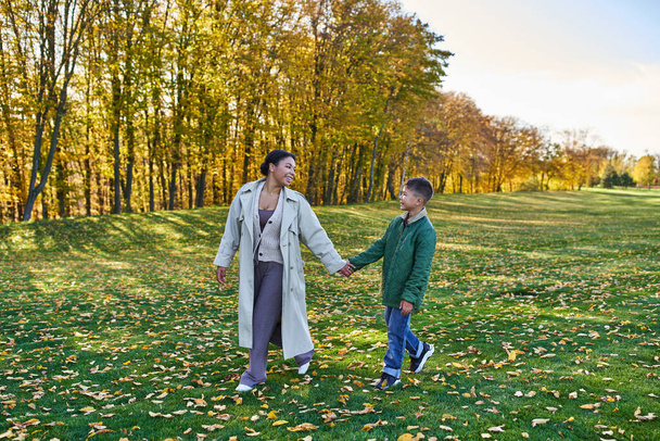 šťastná africká Američanka držící se za ruce se synem, kráčející po trávě se zlatými listy, podzim - Fotografie, Obrázek