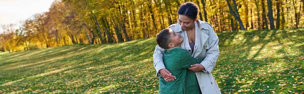 幸せなアフリカ系アメリカ人女性が息子と抱き合って,黄金の葉,秋,旗で草の上に立って - 写真・画像