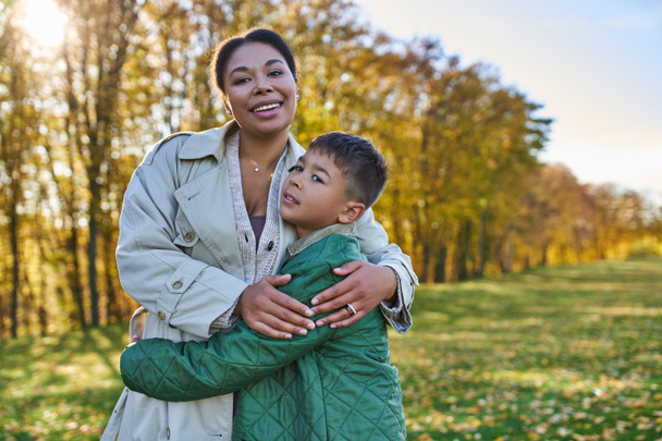 amor maternal, alegría, mujer afroamericana abrazando a lindo hijo, de pie cerca de hojas doradas, otoño - Foto, Imagen