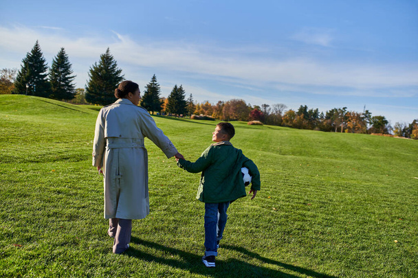 ragazzo afroamericano che tiene il calcio, cammina con la mamma sul campo verde, si tiene per mano, stagione autunnale - Foto, immagini