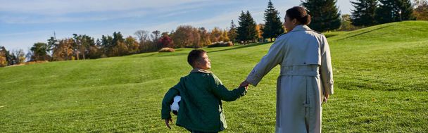 サッカーを保持するアフリカ系アメリカ人の男の子, グリーンフィールドでママと一緒に歩く, 手を握る, 秋, バナー - 写真・画像