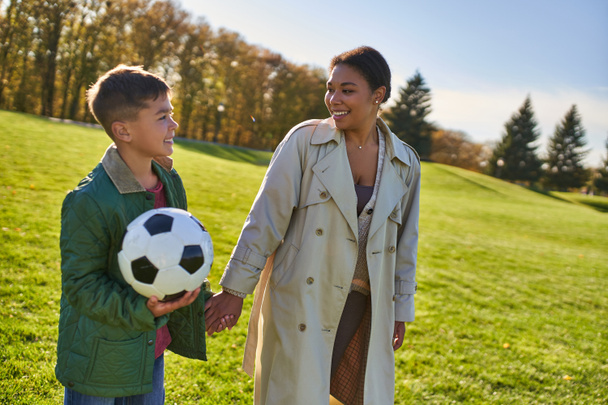 felice ragazzo afroamericano che tiene il pallone da calcio, camminando con la mamma sul campo verde, capispalla, autunno - Foto, immagini