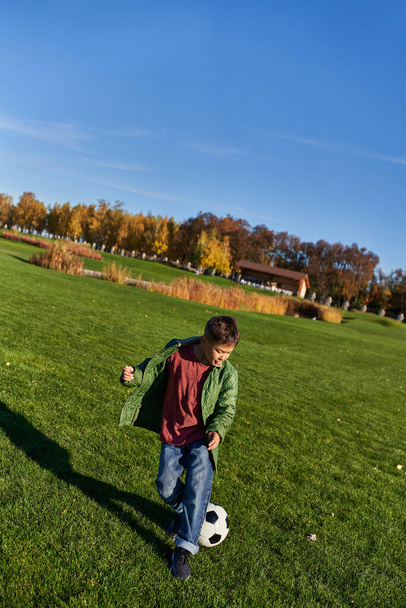 sporty african american boy in autumnal outerwear playing football on green field, soccer, autumn - Photo, Image