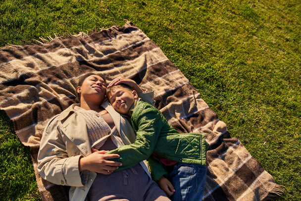 top view, geluk, moederlijke liefde, afrikaanse amerikaanse vrouw en zoon liggend op deken, herfst, gras - Foto, afbeelding
