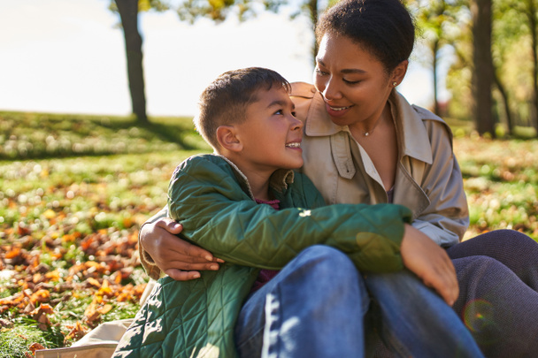 mütterliche Liebe, fröhliche afrikanisch-amerikanische Frau und Sohn umarmen und sitzen neben goldenen Blättern, Herbst - Foto, Bild