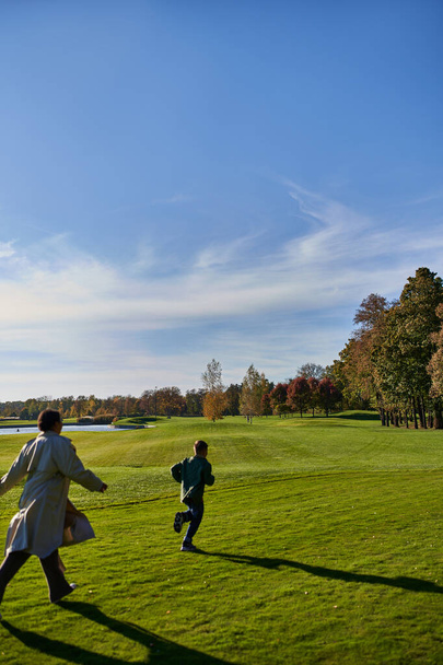 candid shot, african american mother and son running on green grass, having fun, autumn outerwear - Photo, Image