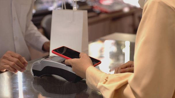 Cep telefonu kullanan üst düzey bir Asyalı Müslüman kadın. NFC kimlik doğrulama ve kimlik doğrulama ödemesi ile birlikte bir satış terminalinde ürün almak için ukalalık yapıyor. - Fotoğraf, Görsel