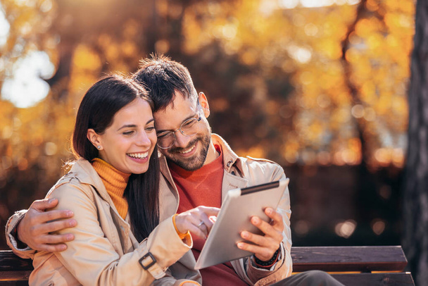 Casal jovem olhando para seu tablet. Eles estão sentados em um banco em um dia ensolarado de outono no parque colorido - Foto, Imagem