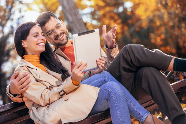 Junges Paar blickt auf sein Tablet. Sie sitzen auf einer Bank an einem sonnigen Herbsttag im bunten Park - Foto, Bild