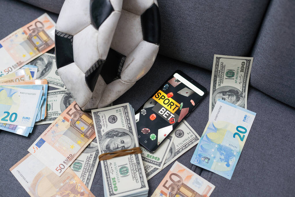 Ставки на спорт онлайн. Доллары падают на фоне руки со смартфоном и футбольным мячом. Творческий фон, азартные игры - Фото, изображение
