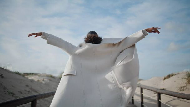 Девушка раздвинула руки на ветреном пляже с видом сзади. Расслабленная женщина наслаждается моментом ощущения свободы на деревянном пирсе. Неизвестный африканский американец отдыхает в выходные. Счастливая женщина в отпуске - Фото, изображение