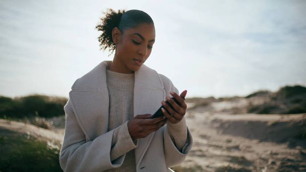 Calm girl message smartphone in sunlight. Serene african american tourist check email social media at beach trip. Beautiful smiling businesswoman work remotely rest seashore. Travel weekend concept. - Foto, Imagen