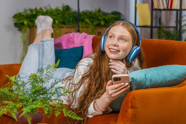 Teenager happy overjoyed child girl in wireless headphones relaxing lying on couch at home apartment choose listening favorite energetic disco dancing music. People weekend daytime leisure activities - Photo, Image