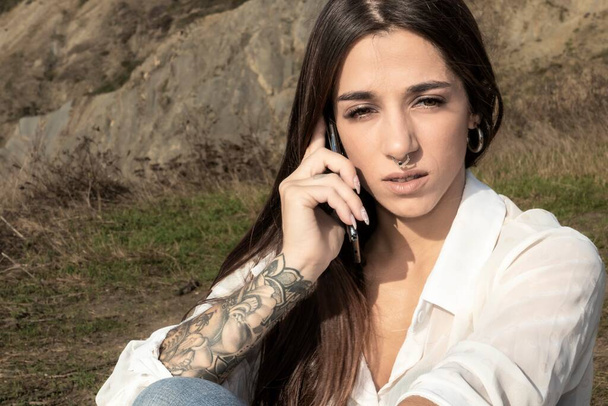Elegante morena con tatuaje de brazo se involucra en conversación telefónica costera - Foto, imagen