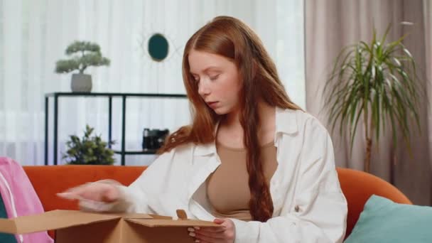 Angry dissatisfied shopper teenager girl unpacking parcel feeling upset and confused with the wrong delivery from an online store, bad quality purchase at home. Adult child indoors in room on couch - Footage, Video