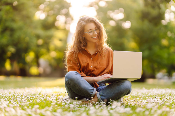 Cute young woman with laptop sits on the grass in the park outdoors. Online training, remote work. The concept of freelance, blog, business. - Foto, immagini