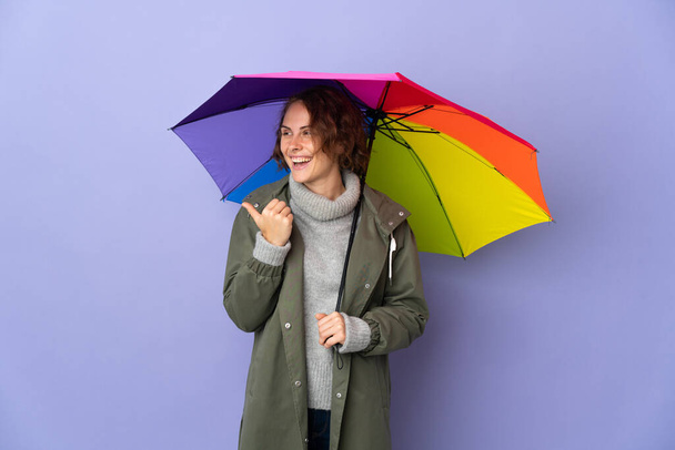 Англійська жінка, що тримає парасольку ізольовану на пурпуровому фоні, вказує на бік, щоб представити продукт - Фото, зображення
