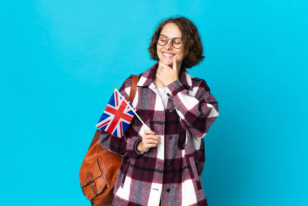 Jonge Engelse vrouw met een vlag van het Verenigd Koninkrijk geïsoleerd op blauwe achtergrond kijken naar de zijkant en glimlachen - Foto, afbeelding