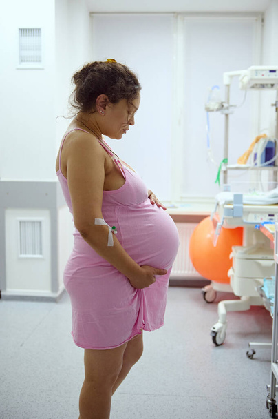 Egy terhes nő oldalsó portréja, a szülőanya tartja a hasát, miközben összehúzódása van a vajúdás alatt, a kórházi kórteremben. Terhesség. Könnyű szülés. Szülés. Szülészet és nőgyógyászat - Fotó, kép
