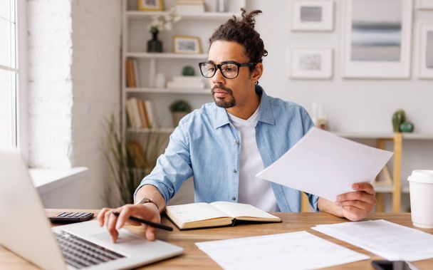 Empresario afroamericano concentrado navegando por netbook y trabajando con documentos mientras está sentado en la mesa en su oficina en el hogar - Foto, Imagen