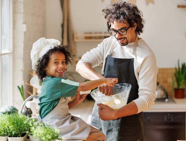 Contenido cariñoso padre afroamericano y lindo niño añadiendo ingredientes en el tazón y preparando la masa en la mesa en casa - Foto, imagen