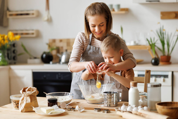 Madre amante del contenido y lindo niño añadiendo ingredientes en el tazón y preparando la masa en la mesa en casa - Foto, Imagen