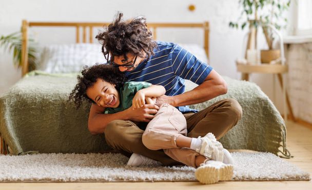 Positiver afroamerikanischer Vater kitzelt lachenden Sohn, während er im Schlafzimmer auf dem Boden sitzt und Spaß hat - Foto, Bild