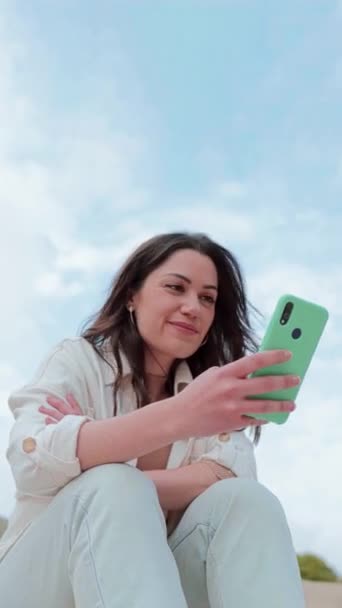 Vertical individual portrait of young adult brunette woman watching content on a cellphone app sitting outdoors. Carefree lady smiling and having fun texting messages with a smart phone. Copy scpace - Footage, Video