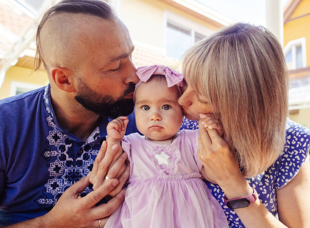 Eltern in ukrainischen Stickereien küssen ihr Kind. Ein kleines Mädchen mit dunklen Augen und ihre liebevolle Mutter und ihr Vater. Das Konzept einer glücklichen Kindheit und die Manifestation von Fürsorge. - Foto, Bild