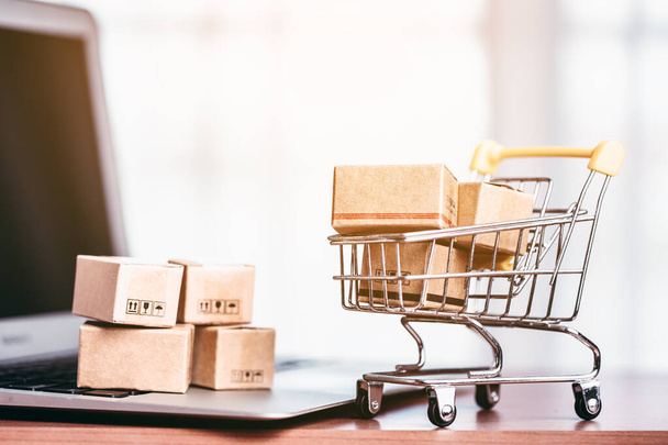 Braune Paketkästen am Laptop und im Warenkorb stehen auf einem Holztisch für das Konzept von Online-Einkauf, Bequemlichkeit und Einkäufen vor der Haustür. - Foto, Bild