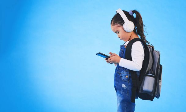 Musiikki, reppu ja lapsi sinisellä taustalla puhelin valmiina kouluun, oppimiseen ja koulutukseen. Lastentarha, studio ja nuori tyttö laukku älypuhelimeen streaming laulu, ääni ja radio. - Valokuva, kuva
