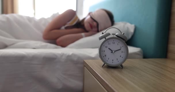 Vintage ξυπνητήρι στέκεται στο ξύλινο τραπέζι κατά κορίτσι σε μάσκα ύπνου κοιμάται στο κρεβάτι σε ελαφριές υποθέσεις. Τεχνολογία για ξύπνημα και όνειρο έννοια αργή κίνηση - Πλάνα, βίντεο