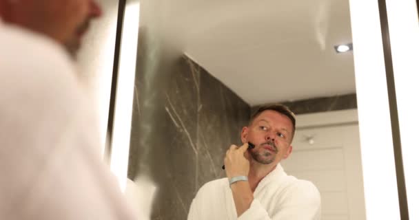 Hombre afeita la barba con afeitadora desechable mirando en el espejo en el cuarto de baño de la vivienda. Concepto de cuidado de la apariencia y rutina diaria en cámara lenta - Metraje, vídeo