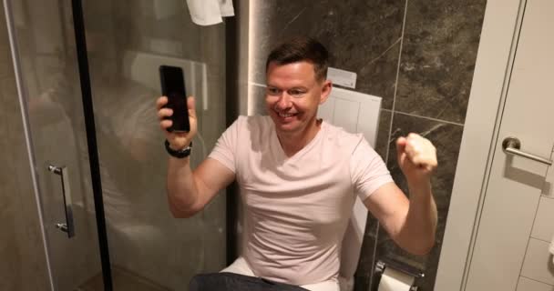 Positive man rejoices in winning game sitting on toilet bowl in bright bathroom of accommodation. Entertainment and daily routine concept slow motion - Footage, Video