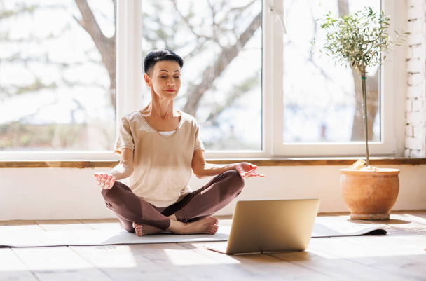 Босонога жінка середнього віку в активному одязі закриває очі і роздумує в позі Лотоса проти вікна під час онлайн-сеансу йоги вранці вдома - Фото, зображення