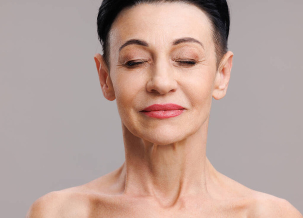 Starší žena 'tvář se stylovým make-up zavírající oči a usmívající se během krásy rutiny na šedém pozadí - Fotografie, Obrázek