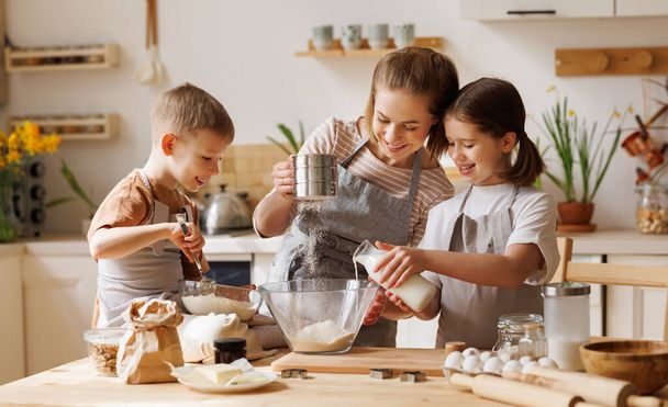 Content loving mother and cute kids: son and daughter adding ingredients in bowl and preparing dough at table at home - Photo, Image