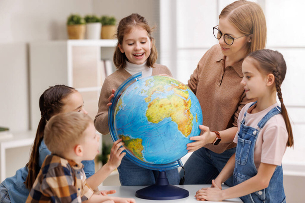 Szemüveges tanár és pozitív gyerekek, akik a földgömböt vizsgálják, miközben az asztalnál állnak a földrajz órán az iskolában. - Fotó, kép