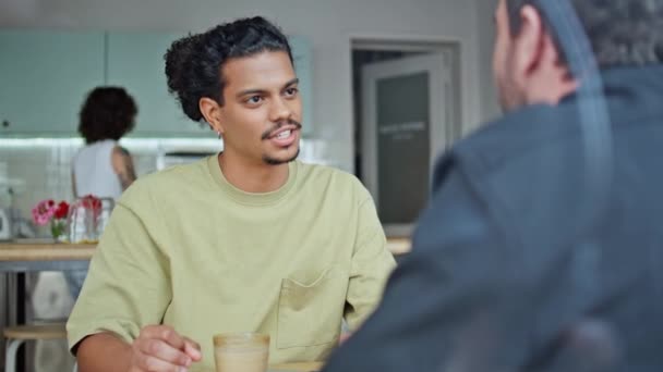 Twee mannelijke vrienden praten in een klein rustig café van dichtbij. Jonge multiraciale mannen die samen praten zitten gezellig koffiehuis. Knappe latijns-amerikaanse man glimlachend naar onbekende gesprekspartner in cafetaria. - Video