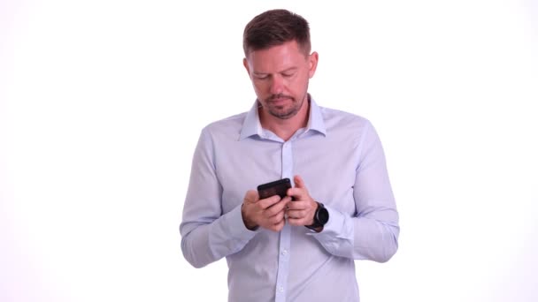 Бородатый бизнесмен просматривает социальные сети на черном смартфоне у белой стены студии. Предприниматель печатает сообщения парню с забавным выражением - Кадры, видео