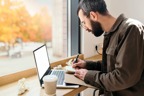 Γενειοφόρος επιχειρηματίας σε casual στυλ γραφής σε ένα σημειωματάριο σε ένα καφέ. εργασία διαδικασία, Ένας άνθρωπος κάθεται σε ένα καφέ με ένα φορητό υπολογιστή, απομακρυσμένη εργασία, freelance, φθινόπωρο - Φωτογραφία, εικόνα