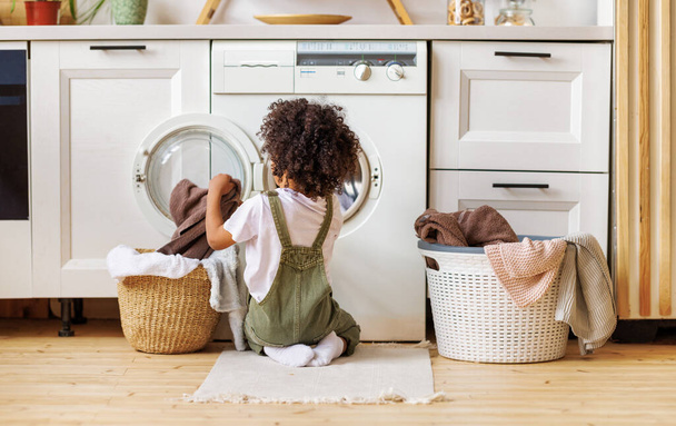 Back view of black boy with curly hair kneeling on floor and putting clothes into washing machine while doing laundry in kitchen at home - 写真・画像