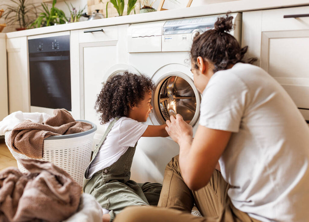 Vidám fekete család: gyerek fia segít apa ágynemű kosárban, miközben csinál mosás közelében mosógép repülés konyha hétvégén otthon   - Fotó, kép