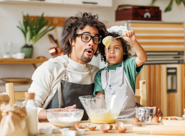 Surpreendido afro-americano homem e menino em aventais preparando massa em tigela enquanto cozinhando juntos à mesa na cozinha - Foto, Imagem