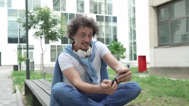 jeden kaukaski człowiek siedzieć na zewnątrz wziąć hamulec szczęśliwy uśmiech grać w gry wideo rozrywka zabawy trzymać telefon komórkowy używać smartfona bawić online dorosły mężczyzna powolny ruch - Materiał filmowy, wideo