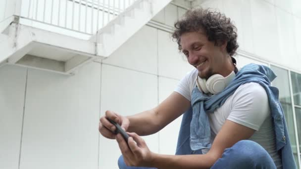 Один кавказький чоловік сидить на відкритому повітрі взяти гальма щаслива посмішка грати в відеоігри дозвілля заняття весело провести мобільний телефон використовуйте смартфон розважайтеся онлайн дорослий чоловік повільний рух - Кадри, відео